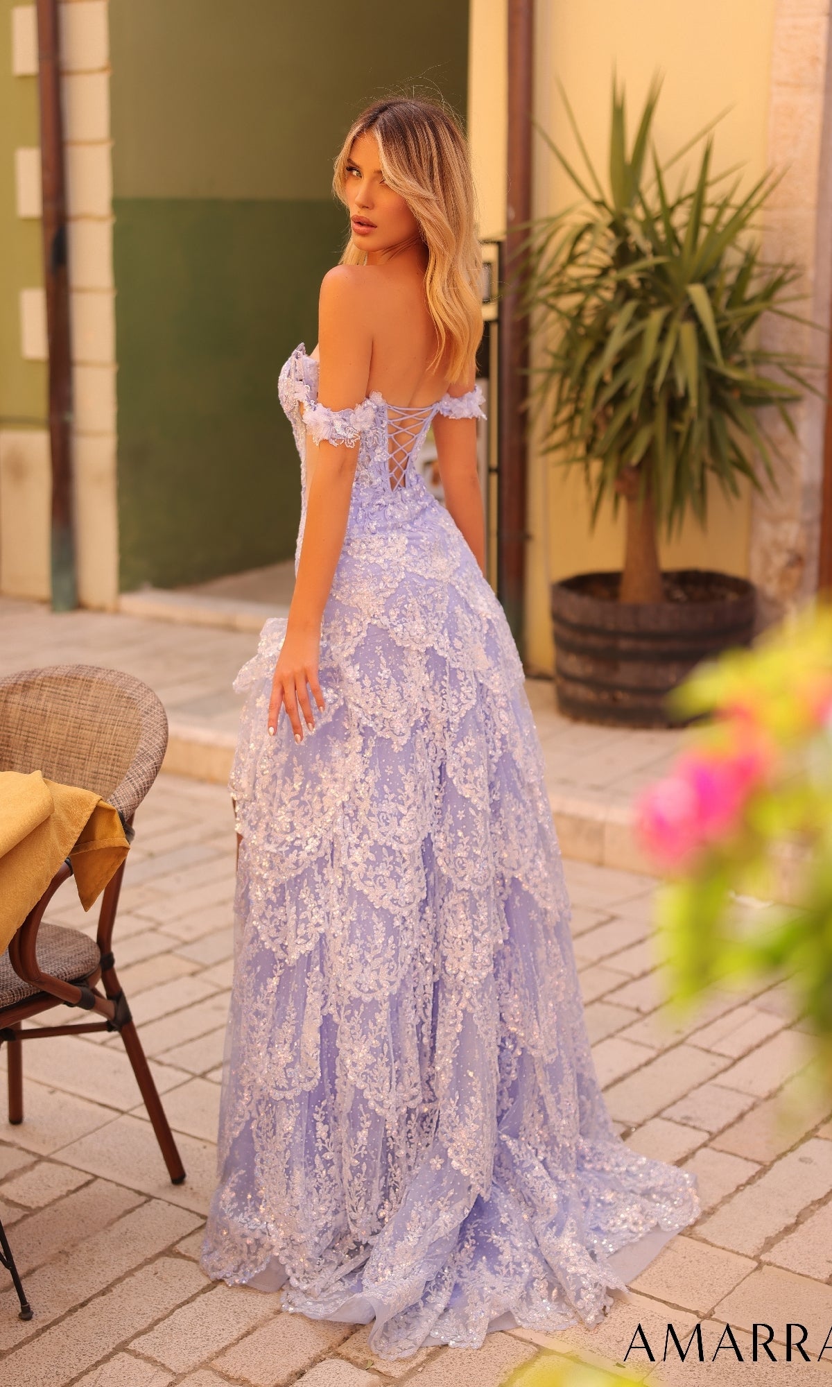 A woman stands outdoors in the Amarra 88251 long prom dress, featuring light purple lace, an off-the-shoulder design, lace-up open back, and a layered skirt. She faces away, glancing over her shoulder.